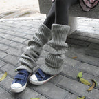 Person wearing grey ribbed-knit leg warmers over dark leggings and blue sneakers on a paved outdoor walkway.