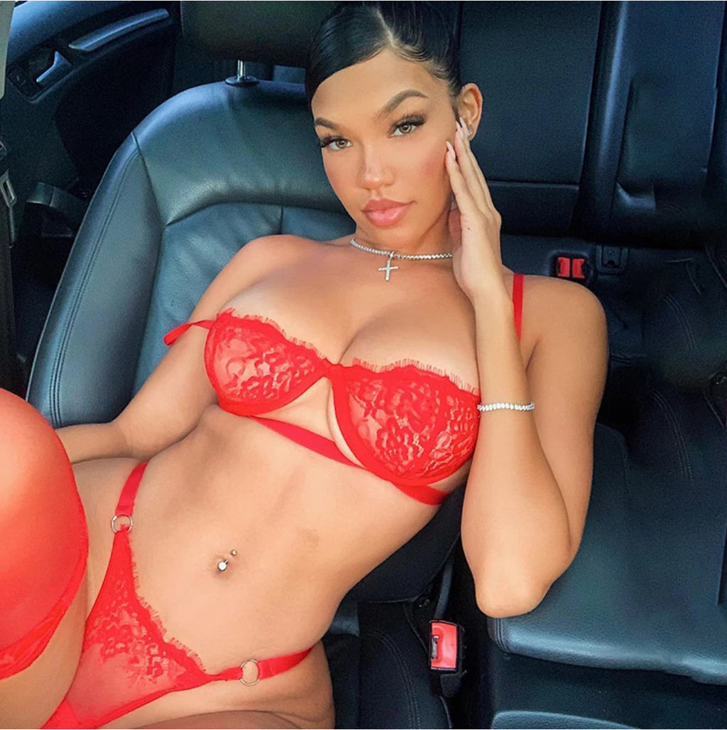 A woman in a red lace underwire bra and matching thong with silver ring details, sitting in a car.