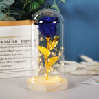 Sparkling blue rose with gold stem and leaves in a glass dome with LED string lights on a wooden base.