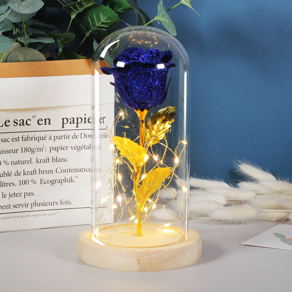 Sparkling blue rose with gold stem and leaves in a glass dome with LED string lights on a wooden base.