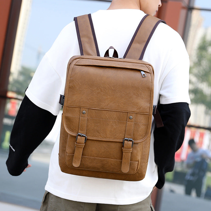 A man wears a tan faux leather retro backpack with buckled straps, a top zipper, and padded shoulder straps.