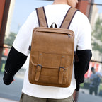 A man wears a tan faux leather retro backpack with buckled straps, a top zipper, and padded shoulder straps.