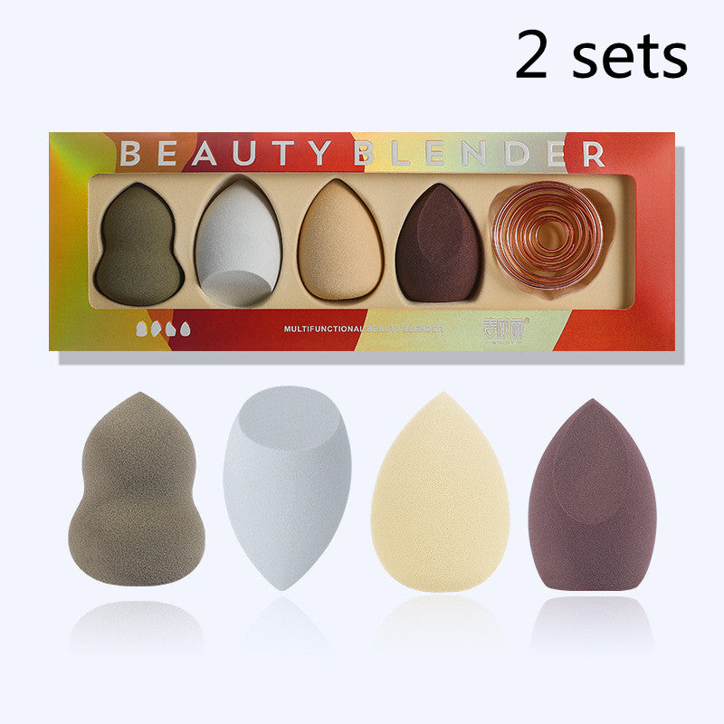 Two sets of four multi-shaped, multicolored makeup sponges with a circular gold metal stand in a branded box.