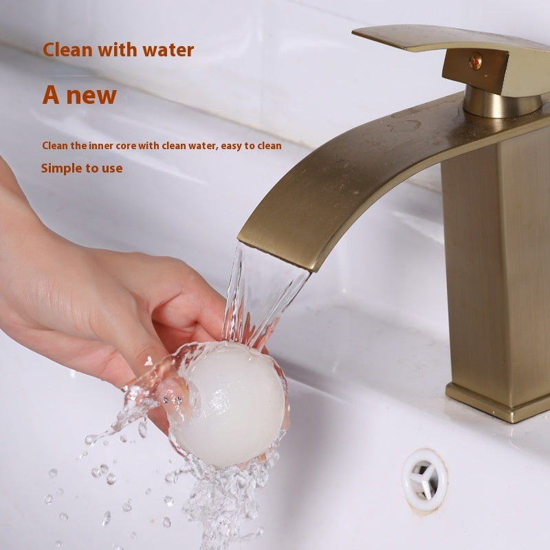 A hand washes a small, translucent, sticky gel hair-removal ball under a modern gold bathroom faucet.