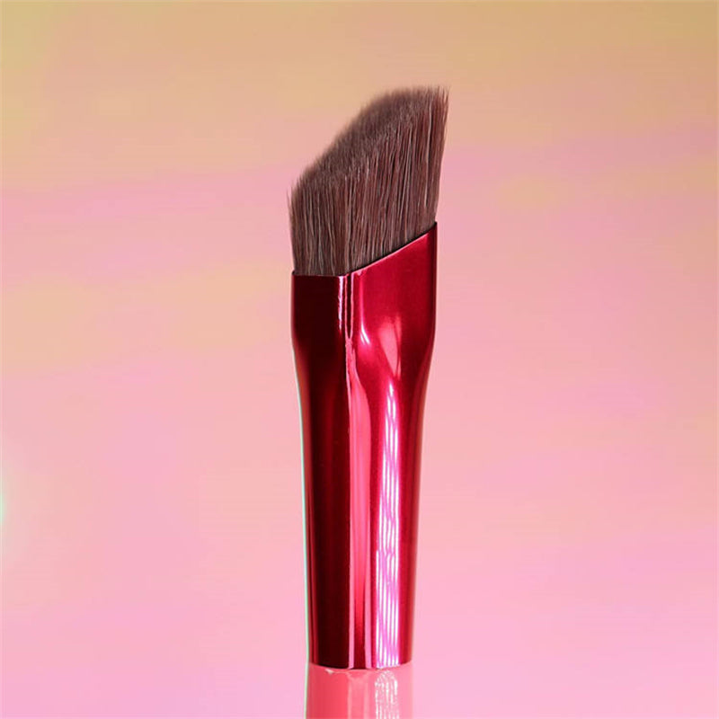 Squared-off angled eyebrow brush with a metallic red handle and soft brown bristles against a peach gradient background.
