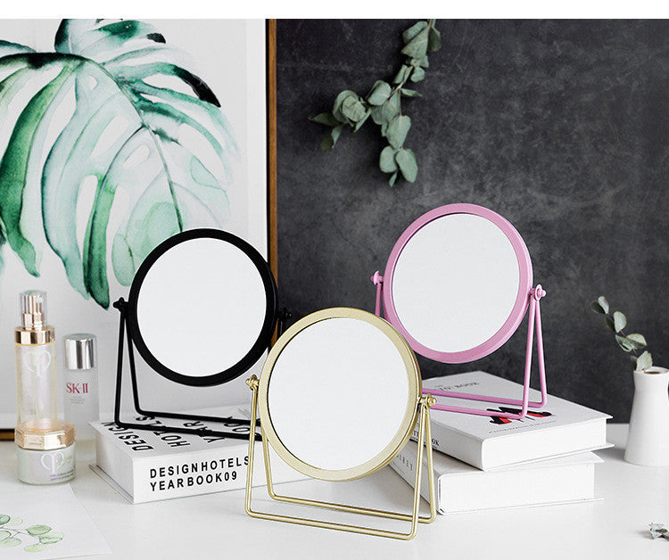 Three round, rotating tabletop makeup mirrors in black, gold, and pink, displayed on books against a decorative backdrop.