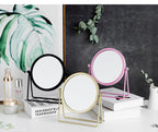 Three round, rotating tabletop makeup mirrors in black, gold, and pink, displayed on books against a decorative backdrop.