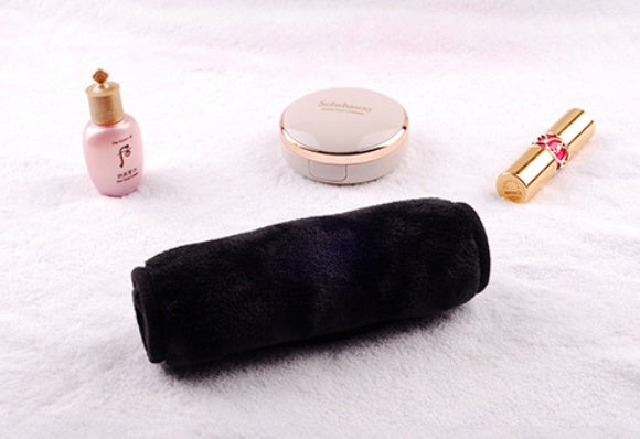 A plush black makeup remover towel on a white surface, accompanied by a small pink bottle, compact, and lipstick.