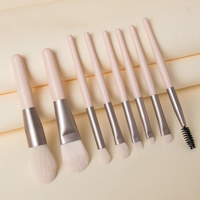 Set of 8 travel makeup brushes with pale pink handles, matte metallic ferrules, and soft white synthetic bristles.