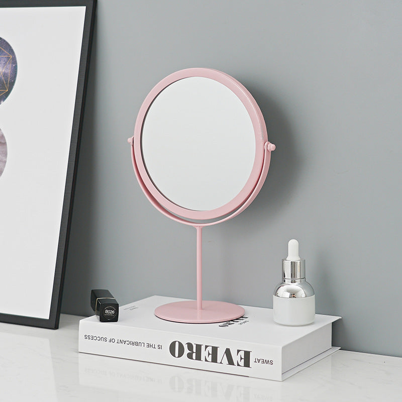 Round pink metal makeup mirror on a pedestal stand, placed on a book against a gray wall.