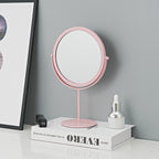 Round pink metal makeup mirror on a pedestal stand, placed on a book against a gray wall.