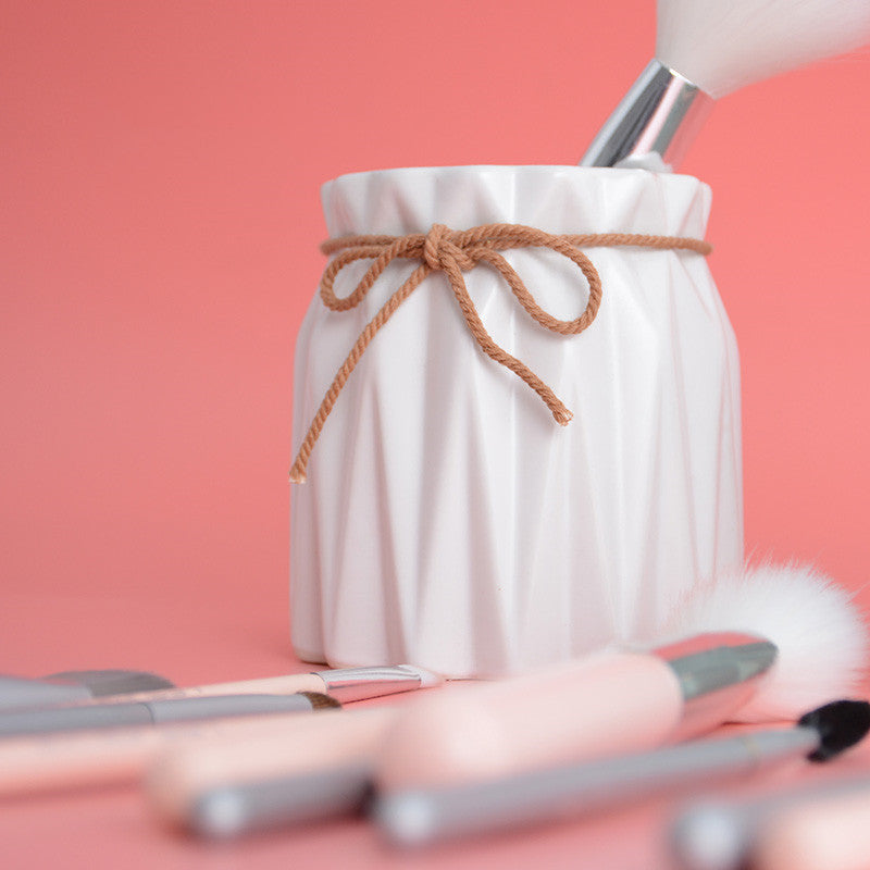 White ceramic, pleated makeup brush holder tied with a rustic twine bow, set against a solid pink background.