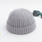 Grey ribbed knit beanie cap with a cuffed brim, shown next to green-framed sunglasses on a white surface.
