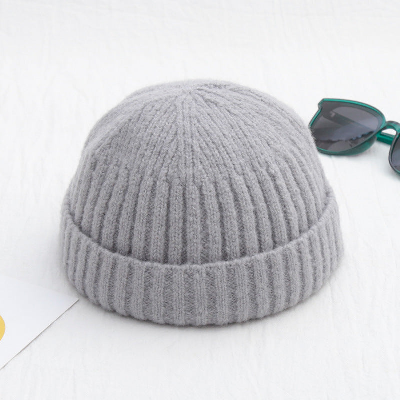 Grey ribbed knit beanie cap with a cuffed brim, shown next to green-framed sunglasses on a white surface.