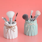 White and mint green fluted ceramic makeup brush holders tied with decorative twine on a pink background.