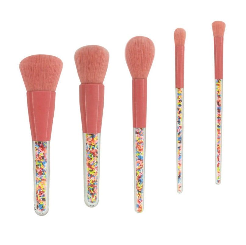 Five makeup brushes with pink bristles, coral ferrules, and clear handles filled with colorful candy-like sprinkles.