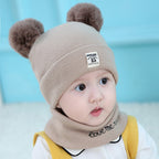 Baby wearing a beige wool hat with two brown faux-fur pom-poms, a logo patch, and a matching neck warmer.