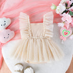 Beige baby jumpsuit with ruffle-trimmed tulle skirt and bodice, displayed on a plush white mat with floral accents.