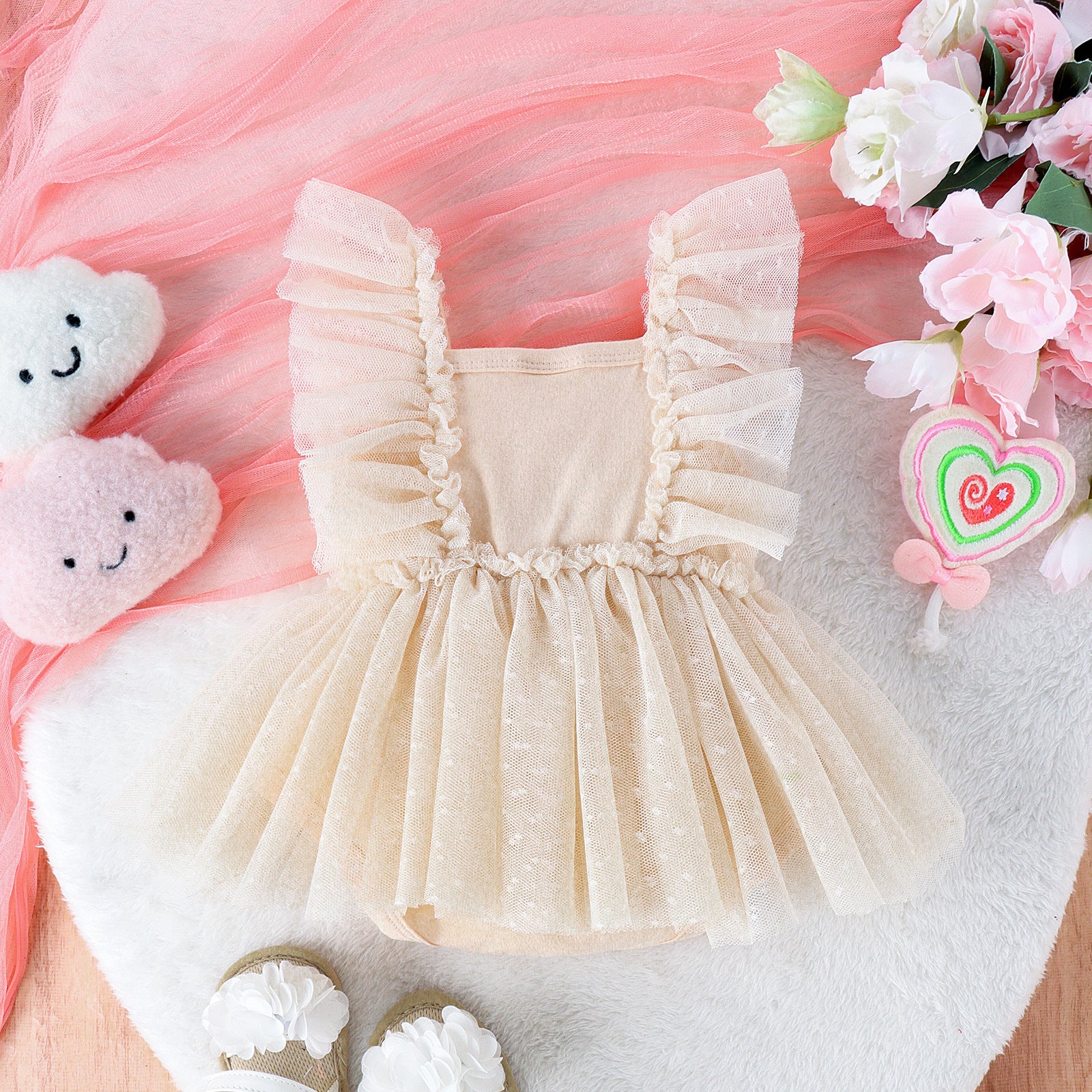 Beige baby jumpsuit with ruffle-trimmed tulle skirt and bodice, displayed on a plush white mat with floral accents.