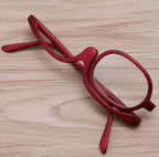 Red makeup reading glasses with a specialized swivel lens frame, lying on a light wood grain surface.