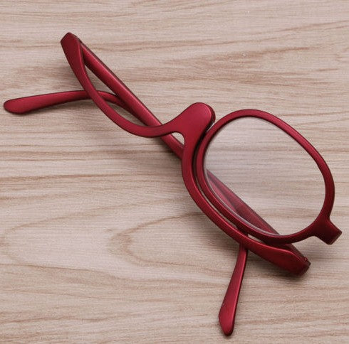Red makeup reading glasses with a specialized swivel lens frame, lying on a light wood grain surface.