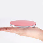 Hand holding a round, pink, portable LED makeup mirror with a metallic silver trim and a USB charging port.