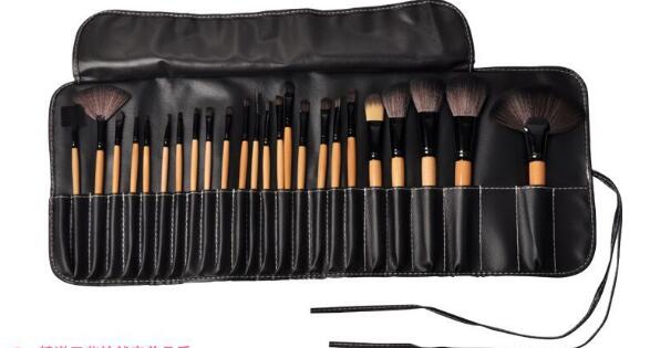 Set of 24 professional makeup brushes with light wood handles and dark bristles, stored in a black leather roll-up bag.