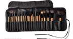 Set of 24 professional makeup brushes with light wood handles and dark bristles, stored in a black leather roll-up bag.