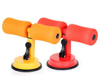 Two sit-up bar trainers with foam padding, metal frames, and suction bases in yellow/orange and red color schemes.