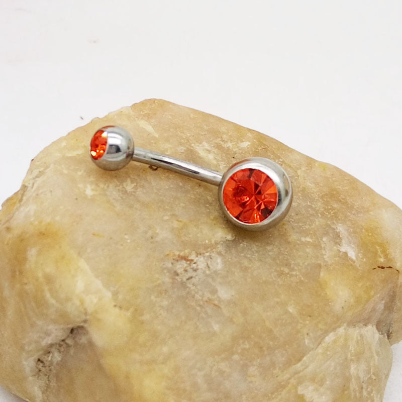 Titanium steel navel barbell featuring two bright orange gemstone settings, resting on a textured tan stone.