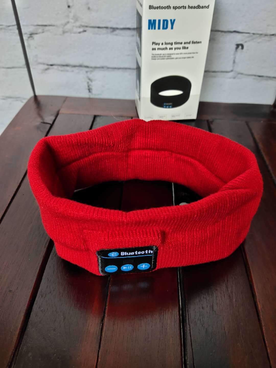Red knitted Bluetooth sports headband with a control panel and matching packaging box on a dark wooden surface.