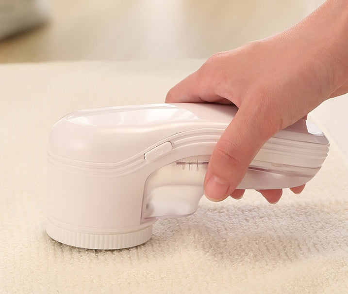 Hand holding a handheld, pale pink electric lint remover on a beige fabric surface.