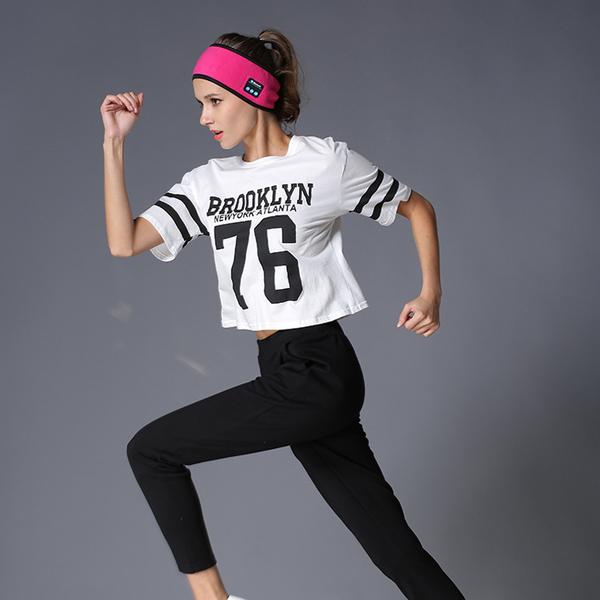 A woman running in a bright pink fabric Bluetooth headband, white athletic jersey, and black pants.