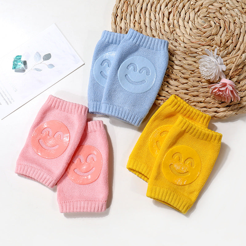 Three pairs of terry cloth baby knee pads in light blue, pink, and yellow, each featuring a smiley face silicone grip.