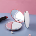 Pink and white portable LED compact mirror with a reflective surface, styled with lipstick and pearls on a blue base.