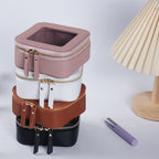 Stacked small square cosmetic bags in pink, white, brown, and black PU leather with transparent tops and gold zippers.