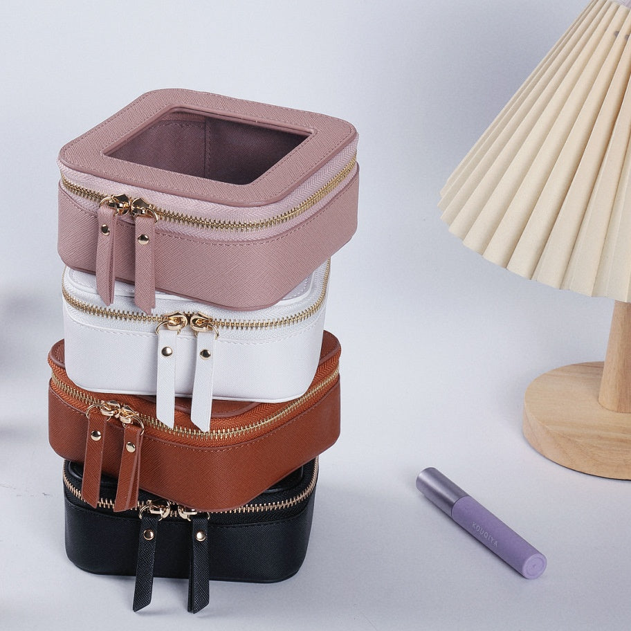 Stacked small square cosmetic bags in pink, white, brown, and black PU leather with transparent tops and gold zippers.