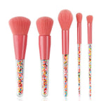 Five makeup brushes with soft pink bristles, matching ferrules, and clear handles filled with colorful candy sprinkles.