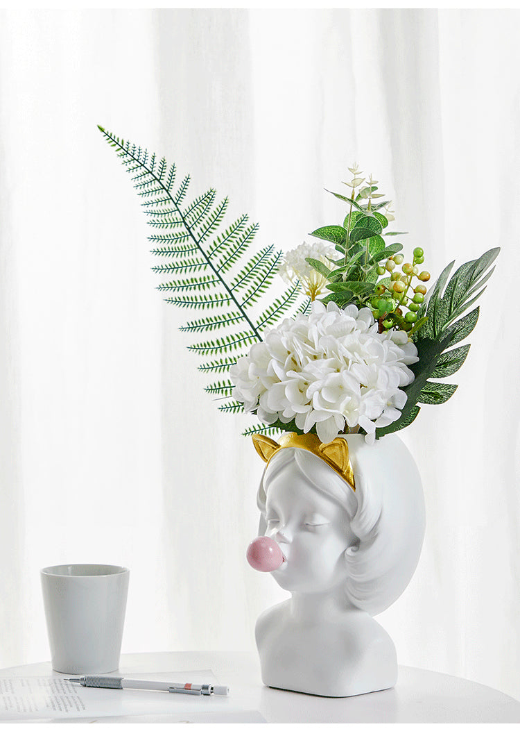 White ceramic girl head vase with a gold cat-ear headband and a pink bubble gum sphere, holding floral arrangements.
