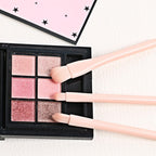 Three light pink makeup brushes resting on a black six-pan eyeshadow palette with shimmering pink and brown tones.