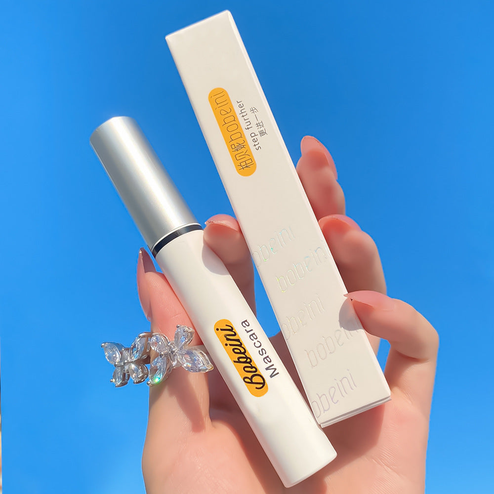 A hand holding a white Bobeini mascara tube and its box against a bright blue sky.