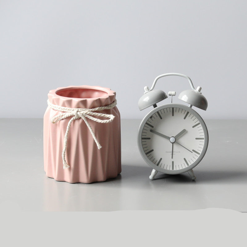 A pink pleated ceramic brush barrel tied with a white string, sitting next to a matching grey twin-bell alarm clock.