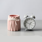A pink pleated ceramic brush barrel tied with a white string, sitting next to a matching grey twin-bell alarm clock.