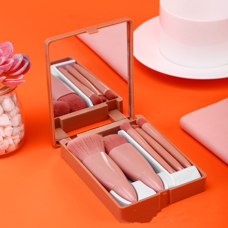 Five-piece pink makeup brush set in a compact case with a mirror, arranged on an orange surface.