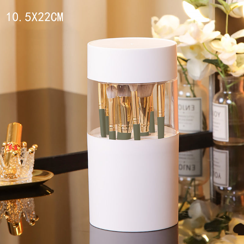 White cylindrical makeup brush storage box with clear center, holding brushes with gold handles and olive green bases.