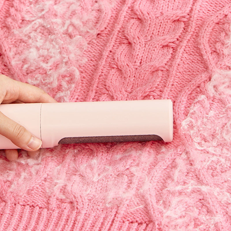 A hand holds a light pink, cylindrical lint remover brush against a pink cable-knit sweater covered in lint and fur.