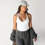 A woman wears a white, ribbed-knit racerback tank top with a V-neckline, styled with a cap and gray sweats.