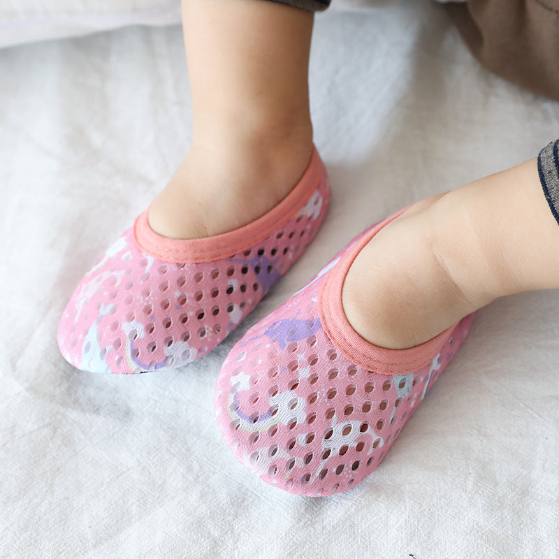 Baby feet wearing pink mesh first-step shoes with unicorn prints and soft pink trim on a white fabric background.