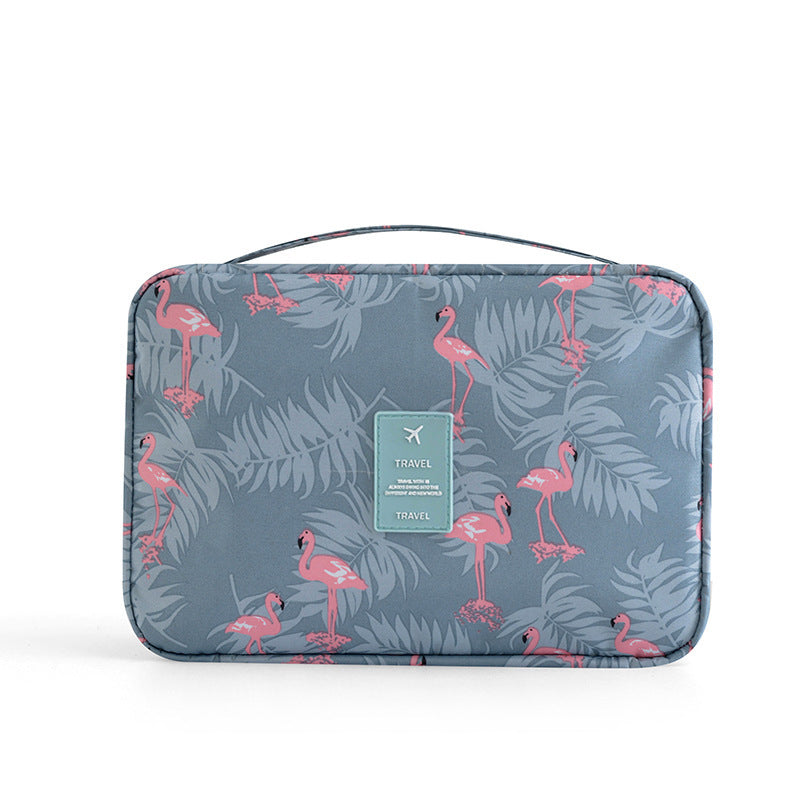 Blue travel makeup bag with pink flamingo and grey tropical leaf print, top handle, and teal "TRAVEL" brand patch.
