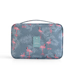 Blue travel makeup bag with pink flamingo and grey tropical leaf print, top handle, and teal "TRAVEL" brand patch.